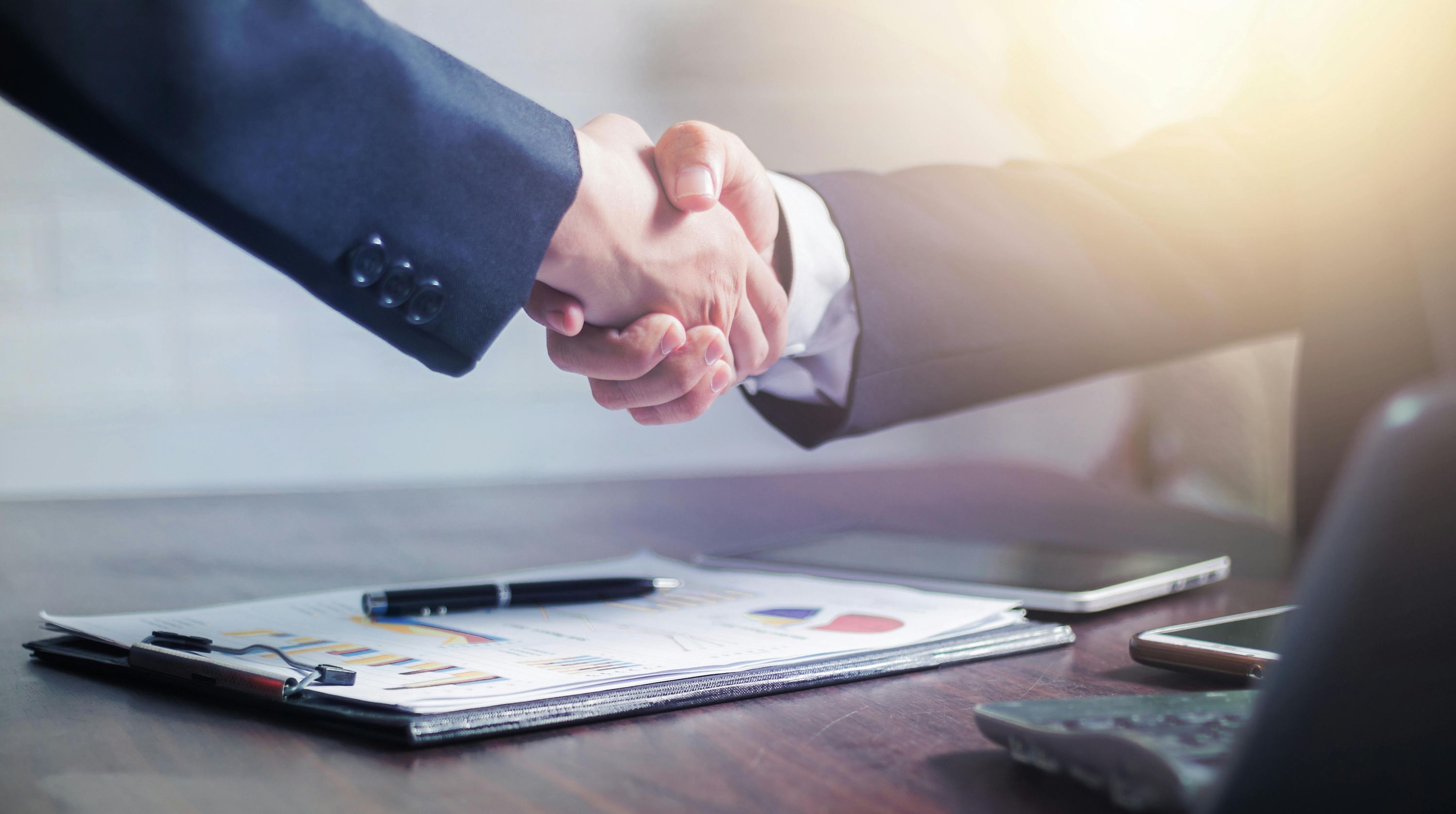 Two businesspeople shaking hands over a desk with charts and documents
