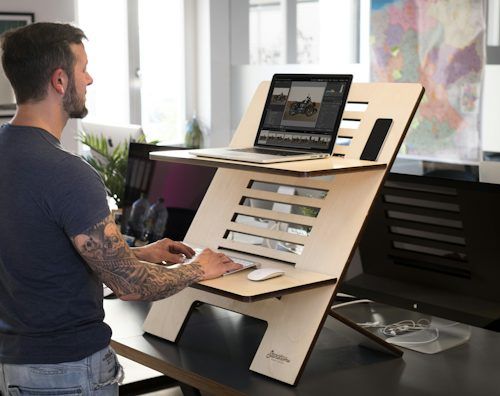Man working on a laptop at a wooden standing desk in a bright office