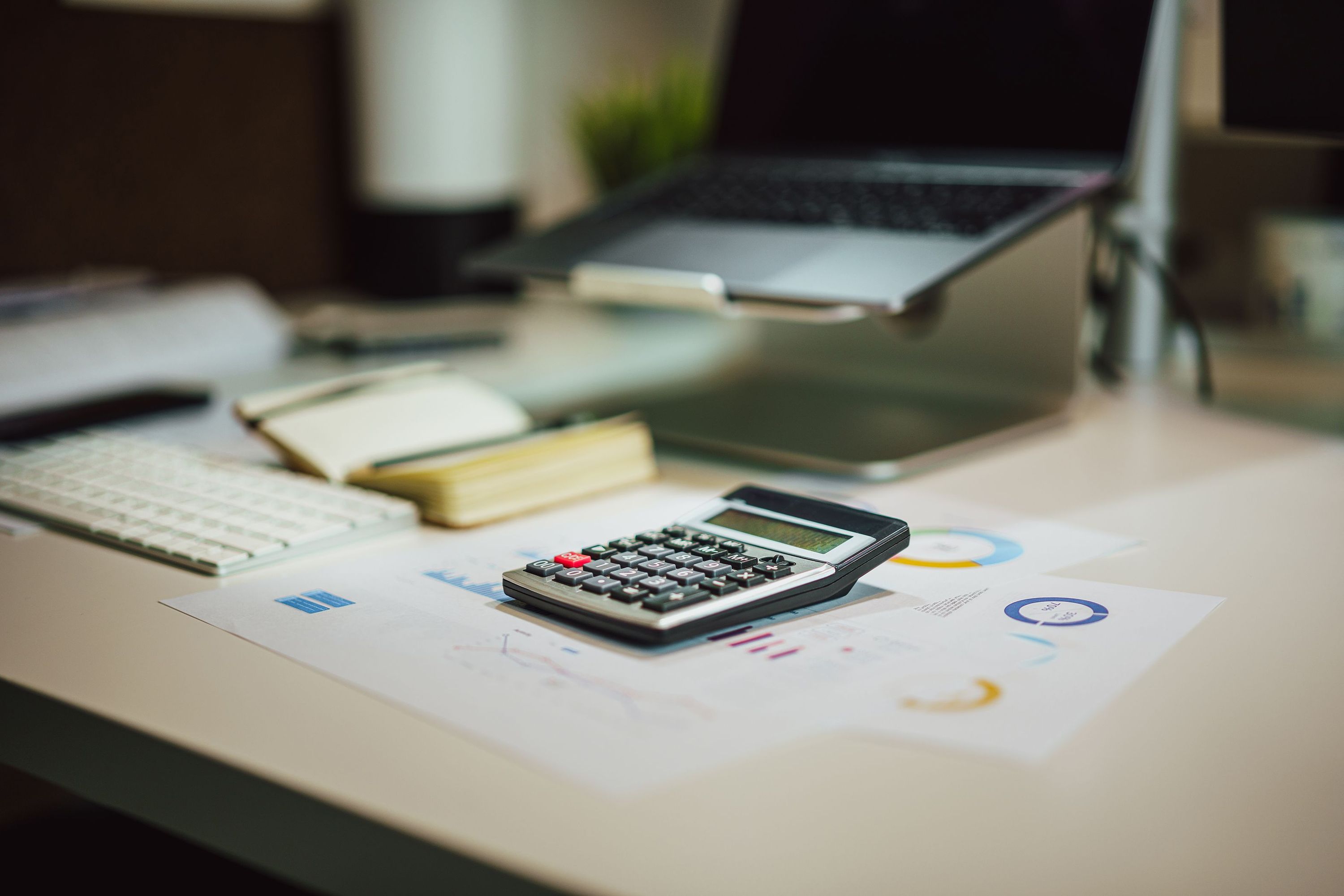 Calculator on printed charts beside a keyboard, notepad, and laptop on a desk