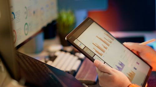 Person reviewing charts on a tablet beside a laptop and desktop monitor