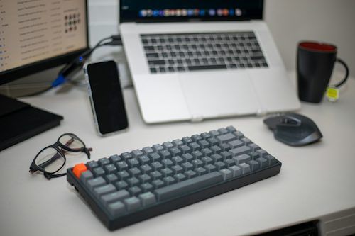 Keyboard and laptop on a desk with a monitor, phone, glasses, and mug