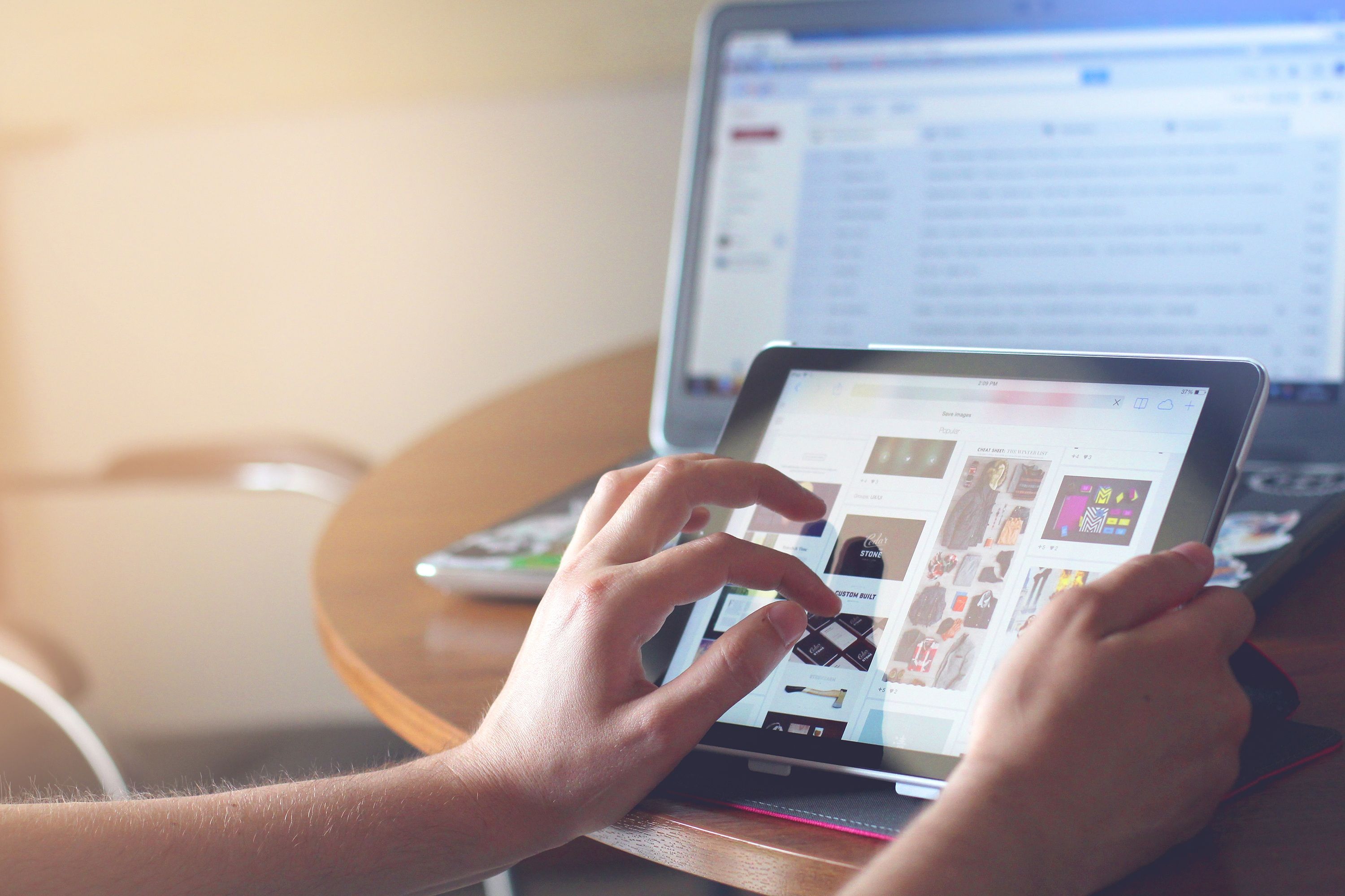 Person browsing a tablet beside an open laptop at a wooden table